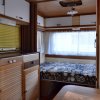 Interno di una roulotte vintage con letto, scaffali in legno e tende bianche.