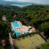 Veduta aerea di piscine, campi da tennis e vegetazione rigogliosa vicino al mare.