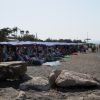 Spiaggia affollata con ombrelloni e lettini, vicino al mare con grandi rocce.
